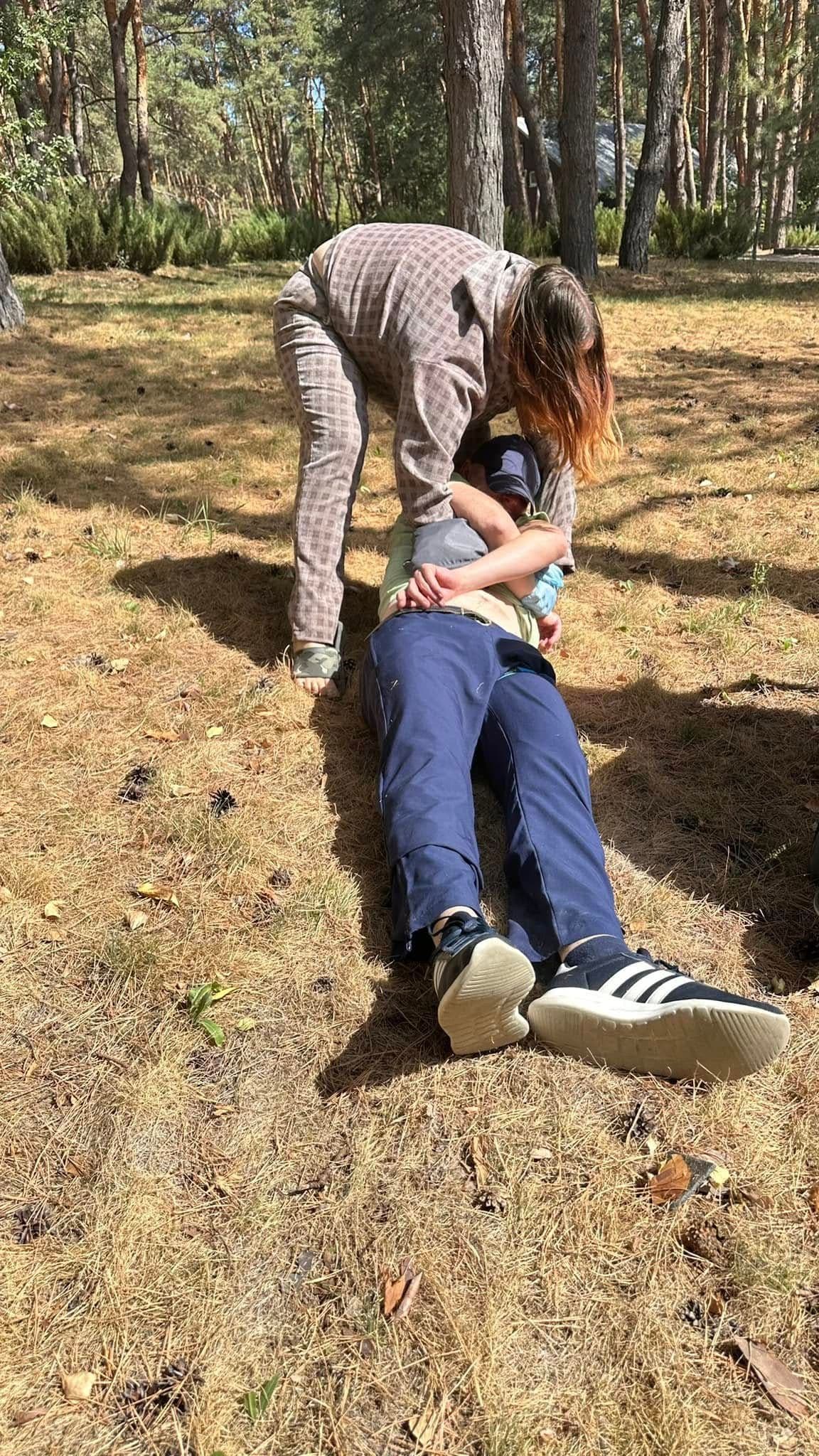 A woman is leaning over a lying man in front of a forest, providing assistance.