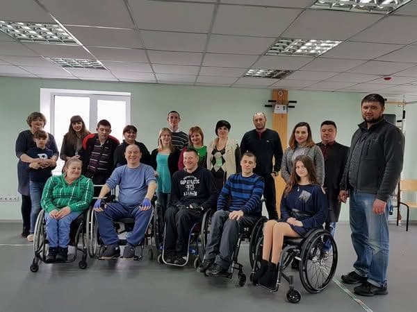 A group photo in a room that looks like a rehabilitation center. In the center, five people in wheelchairs are surrounded by standing participants, including men, women, and even a child. Everyone looks friendly, with many smiling. The room contains exercise equipment, and a window lets in natural light. The atmosphere in the photo feels warm and supportive.